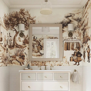 A brown-toned countryside wallpaper with illustrated villagers, animals, and trees, applied in a bathroom with a vanity mirror and sconces.