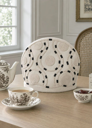 The Cozy Trace tea cosy in Ivory, view-diningroom beside a patterned teapot and jam bowl.