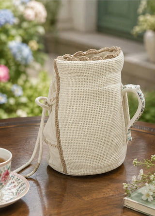 Front view of the Deep Lore ivory jug cover showing the linen tie bow and ladder-stitch panels over a floral teapot in a light dining room.