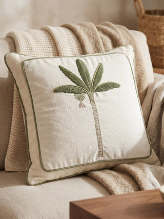 A close-up of a luxury cream cushion featuring a hand-embroidered sage green palm tree on a soft neutral sofa.