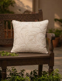 The Eden off-white embroidered linen cushion cover styled on a dark carved wood garden bench surrounded by green foliage.