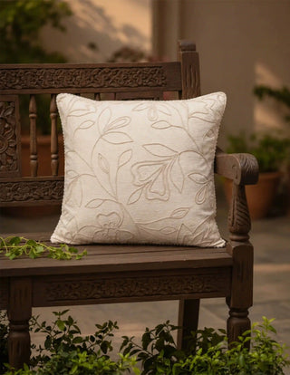 The Eden off-white embroidered linen cushion cover styled on a dark carved wood garden bench surrounded by green foliage.