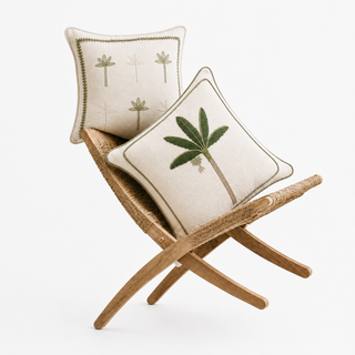The Eka Kanan Botanical Stitch cushion set styled on a woven rattan bench, showing a scattered palm motif cushion with a leaf-vine border and a banana leaf tree cushion, both embroidered in olive on natural cotton-linen.