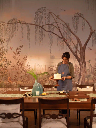 The Enchanted Grove wallpaper mural, with a weeping willow and birds, on a dining room feature wall behind a dark wood table and chairs. view-dining
