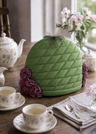 The Finch & Flower tea cosy in Moss & Mulberry styled on a wooden table with a floral teapot, teacups and fresh flowers.