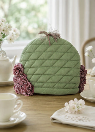 The Finch & Flower tea cosy in Moss & Mulberry on a table with white teacups, a teapot and fresh white flowers.