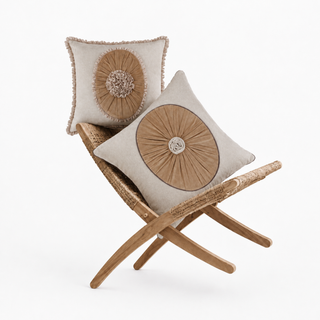 The Florence Pleated & Frieze cushion set styled on a woven rattan bench, showing a ruffled-edge cushion with an oval pleated sunburst medallion and gathered rosette, and a piped cushion with a round pleated sunburst medallion and rolled rosette, both on natural cotton-linen.