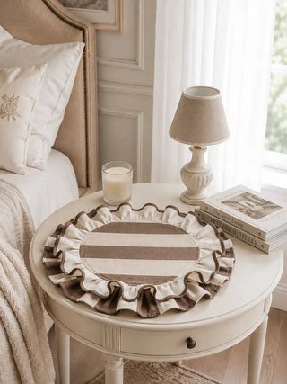 Florentine Ruffles beige and taupe striped table mats with hand-stitched scalloped ruffles styled on a white round nightstand with cream candle, books, and soft bedroom light.