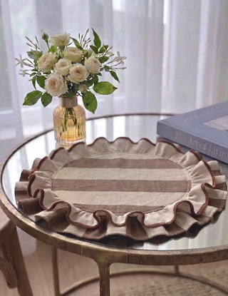 Florentine Ruffles round table mat in beige and mocha placed on a glass-topped brass side table with a glass vase of white roses and a coffee table book in a light-filled room