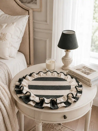 Florentine Ruffles dark green and cream striped table mats with hand-stitched scalloped ruffles styled on a white round nightstand with black linen lamp, cream candles, luxury books, and cream bedding in a serene bedroom setting.