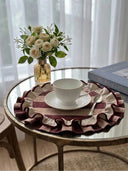 Close-up of Florentine Ruffles mats on a round table with cream teacup, saucer, pale garden roses, and gold-rimmed glassware in soft natural light.