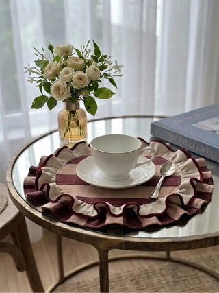 Close-up of Florentine Ruffles mats on a round table with cream teacup, saucer, pale garden roses, and gold-rimmed glassware in soft natural light.