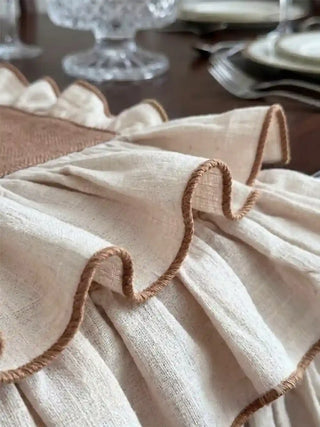 Close-up detail of Florentine Ruffles table runner showing the triple-tier ivory gathered ruffle with fine mocha overlock blanket stitch edge finish, crystal glassware and white plates in soft background focus