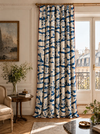 The French Town curtain in Navy & Cream hung from a brass rod in a living room, showing an illustrated townscape of shuttered buildings, cafés and trees against a French-door view.