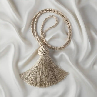 Flat lay of the Frosted Silk Holdback on a satin background, showing the braided cord curled and the full spread of the luminous tassel.