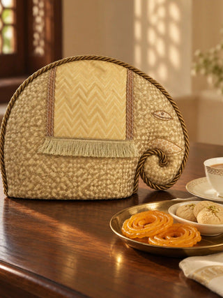 Gajraja elephant tea cosy on a dark wood table beside jalebi and a cup of chai, edged in twisted gold rope with a curled trunk.