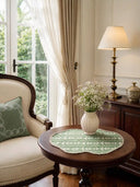 Garden Gala sage green and cream botanical table mats with scalloped borders styled on a dark wood round table with white ceramic vase, baby's breath flowers, brass lamp, and cream wingback chair beside garden windows in natural light.