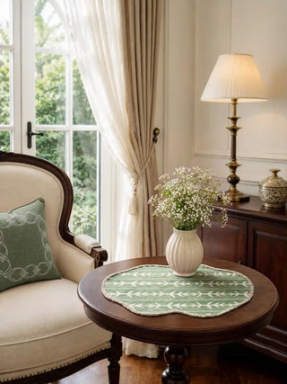 Garden Gala sage green and cream botanical table mats with scalloped borders styled on a dark wood round table with white ceramic vase, baby's breath flowers, brass lamp, and cream wingback chair beside garden windows in natural light.