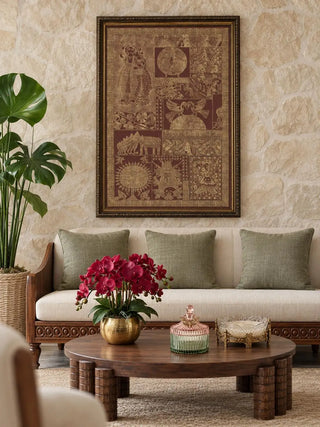 Geet Madhubani embroidered wall art in a dark gold-edged frame on a stone wall above a cream sofa with sage cushions and a carved round coffee table