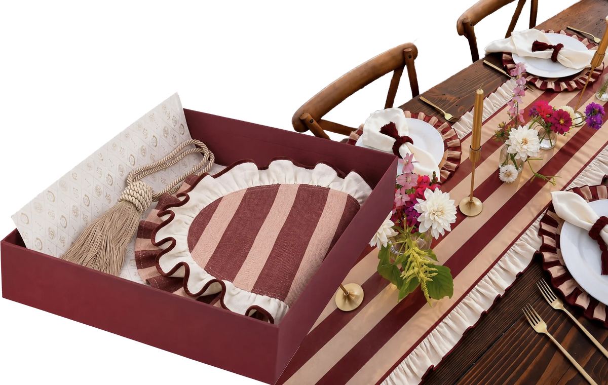 Open gift box of striped ruffled placemats and a tassel tieback beside a dressed dining table