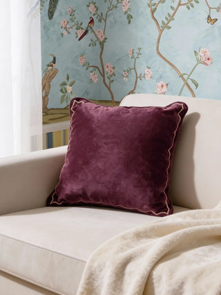 A luxury plum-toned velvet cushion with a gold scalloped embroidered border styled on a white armchair against a botanical mural.