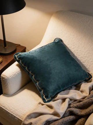 A luxury teal velvet cushion with golden scalloped embroidery styled on a white bouclé armchair next to a black lamp.