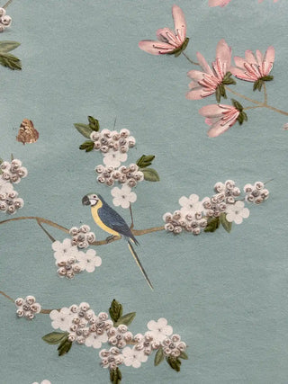 A detailed macro close-up of the Grace Embrace embroidered wall art showing a blue and yellow parrot among white blossoms worked in knotted stitches.