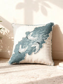 A luxury Graceful Cranes drift cushion featuring teal wave embroidery and white cranes resting on a plush cream rug in a sunlit room.