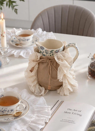 The Grandeur Ruffle jug cover in Taupe, view-diningtable on a floral jug beside a lit candle and teacups.