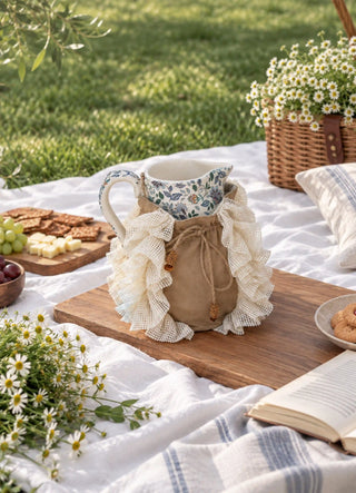 The Grandeur Ruffle jug cover in Taupe, view-picnic on a floral jug beside a wicker basket and daisies.