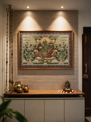 The Green Tara wall art in a wood frame, lit by twin spotlights above a wooden shelf styled with brass pots and offering bowls.