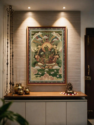 The Green Tara wall art in a wood frame, lit by twin spotlights above a wooden shelf styled with brass pots and offering bowls.