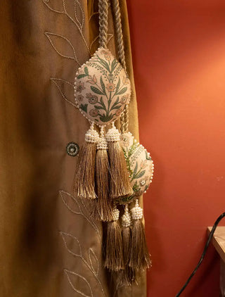The Gulbano tie back in cream with two padded teardrop pods, green floral motif, pearl-like bead edging and gold tassels, on a gold curtain.