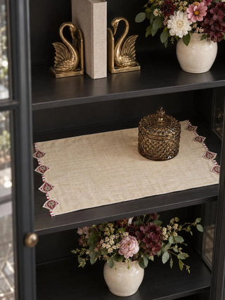 The Gulnaz embroidered table mat in oat with burgundy teardrop motifs and beaded strands, styled on a black cabinet shelf under a glass jar.