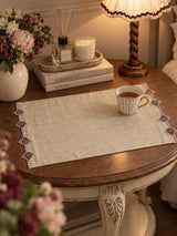 The Gulnaz embroidered table mat in oat with burgundy teardrop motifs and red and white beads, holding a striped teacup on a green table.