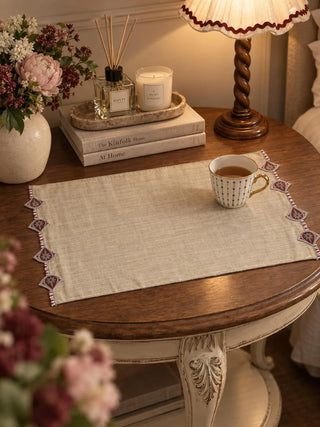 The Gulnaz embroidered table mat in oat with burgundy teardrop motifs and red and white beads, holding a striped teacup on a green table.