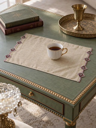The Gulnaz embroidered table mat in oat with burgundy teardrop motifs, on a round wooden side table with a teacup, books and flowers.