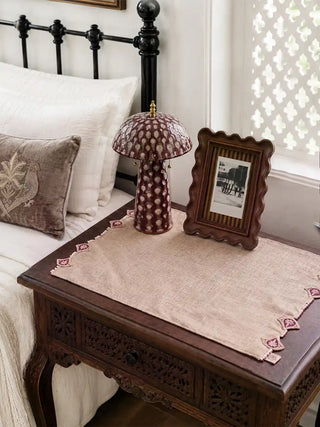 Gulnaz hand-embroidered natural linen table mats with maroon teardrop motifs and pearl bead borders styled on a dark carved wood bedroom console with maroon patterned lamp, framed photograph, and embroidered pillow accent.