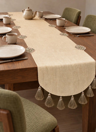Gulnaz table runner in ivory with embroidered floral medallions and a tassel fringe, falling off a wooden table set with stoneware tea cups