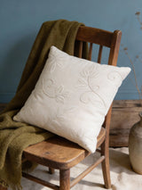 A luxury hand-embroidered pearl cushion cover in ivory with botanical vine details resting on a vintage wooden chair.