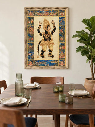 Hanuman Ansh wall art in a whitewashed wooden frame above a walnut dining table set with ribbed green glassware, beside a potted fiddle-leaf fig.