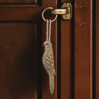The Heer Parrot Gift Charm in Green & Gold, hung from a brass door handle by its braided cord, showing the sequinned body and red beak.