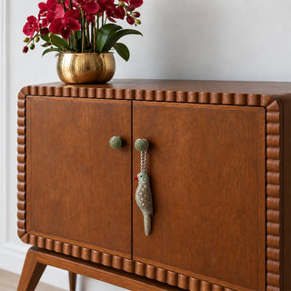 The Heer Parrot Hanging Gift Charm hanging from a green knob on a scalloped-edge wooden cabinet with red orchids in a gold vase.