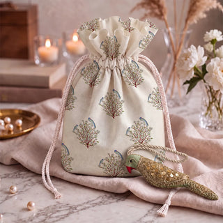 The Heer Parrot Hanging Gift Charm in green and gold resting beside a cream drawstring pouch printed with birds on flowering trees.