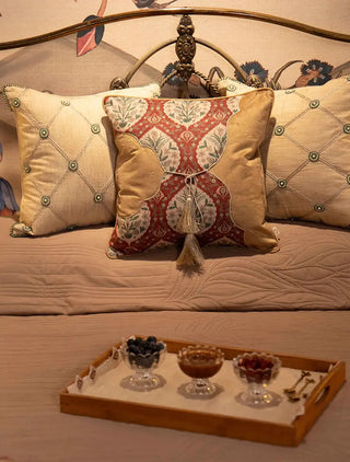 Front view of the Heritage Tie cushion cover in rust with floral medallions, gold scalloped panels and a tasselled cord tie, styled on a taupe quilt.