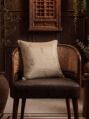 The Hiranya gold embroidered silk cushion cover styled elegantly on a dark leather and cane barrel chair in a wood-paneled room.