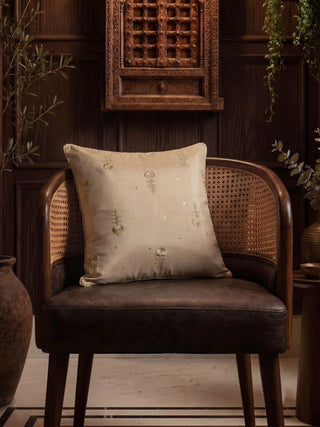 The Hiranya gold embroidered silk cushion cover styled elegantly on a dark leather and cane barrel chair in a wood-paneled room.