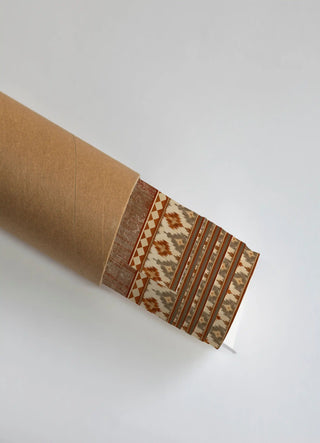 The Ikat Fusion Industrial wall art print partially unrolled from its kraft paper shipping tube, showing a distressed rust-toned texture panel beside an ikat diamond stripe pattern in rust, grey and cream.