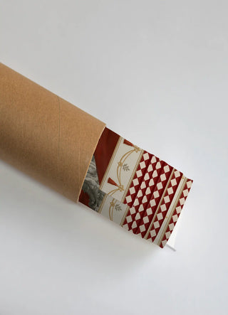 The Invention Modern Fusion wall art print partially unrolled from its kraft paper shipping tube, showing a tasselled garland swag beside a diamond harlequin panel in red, cream and gold.