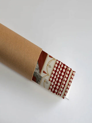 The Invention wall art print partly unrolled from its shipping tube, showing a tasselled garland swag beside a diamond harlequin panel in red, cream and gold.